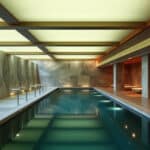 A modern circular sauna with wooden benches lining the walls, a central metal stove filled with rocks, and a gong hanging on the right. Natural light enters from a round ceiling window.