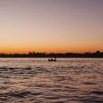 A traditional sailing vessel navigating the Nile River at sunrise, with temple ruins and palm trees visible along the di