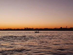 A traditional sailing vessel navigating the Nile River at sunrise, with temple ruins and palm trees visible along the di
